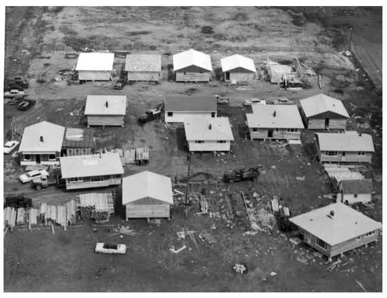 Aerial view of J.B. Ross Construction’s site shows 13 houses in various stages of construction. At far left, movers prepare one home for transport to its new location